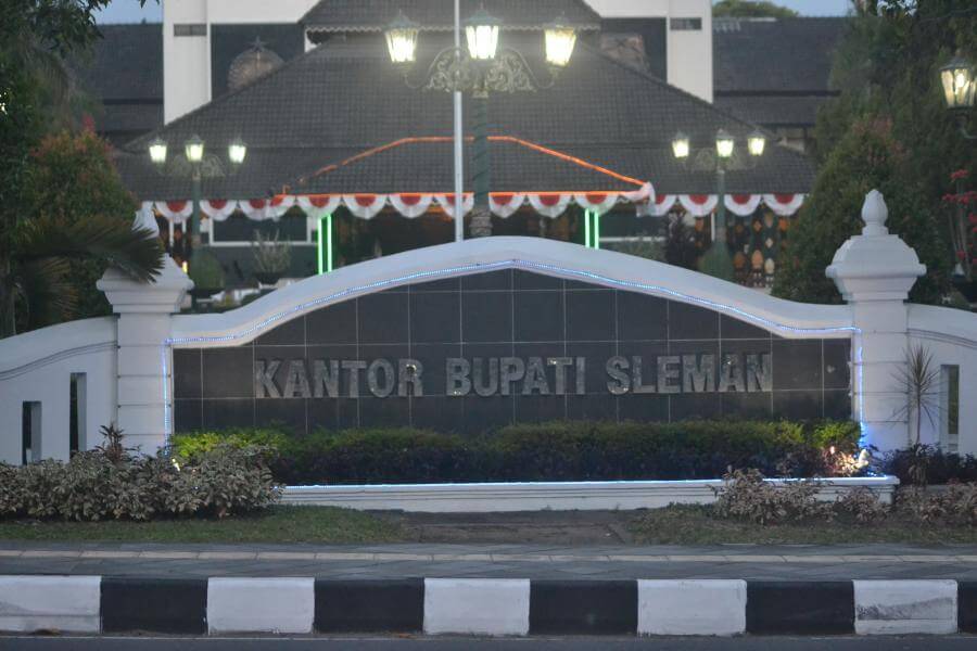 Signage at a gov’t building reading 'KANTOR BUPATI SLEMAN' behind a curved white wall with hedges and lights at dusk/uniformed garden area
