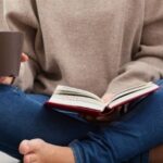 Person sitting cross-legged on the floor in a beige sweater and blue jeans, holding a mug and an open book, with stacks of books nearby for a cozy reading scene.