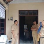 Four people in beige uniforms stand outside a doorway of a small office, chatting near plastic chairs and a signboard mounted on the wall above.