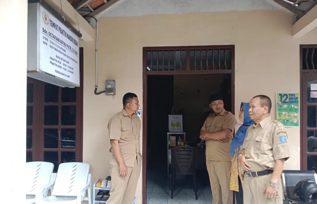 Four people in beige uniforms stand outside a doorway of a small office, chatting near plastic chairs and a signboard mounted on the wall above.