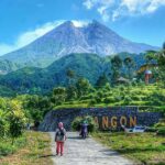 Volcano mountain rising above green hills, with a stone wall sign reading 'ANGON' and people walking along a path.