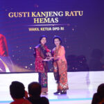 Two women in colorful batik on a lit stage exchange an award, with a large backdrop displaying a portrait and Indonesian text behind them.