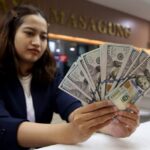 Woman in a blazer at a bank counter fanning out multiple US hundred-dollar bills in her hands.