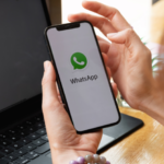 Hand holding a smartphone displaying the WhatsApp app screen beside a laptop.