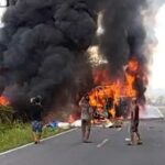 Bus on fire in the middle of a road, large black smoke plumes rising as bystanders watch nearby