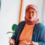 Woman in a pink hijab and glasses speaking while presenting, with hands gesturing near a monitor in a bright room.