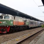 Diesel locomotive in green, white, and orange pulling a passenger train at a platform station with a shelter overhead.