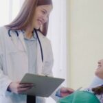 Female doctor in a white coat with a stethoscope and clipboard talking to a patient in a hospital bed.