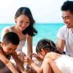Family of four at the beach, kneeling in the sand and playing with sand near the turquoise sea, smiling and engaging together.