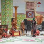 Group of musicians in traditional attire performing gamelan on an outdoor stage with colorful fabric panels behind them