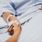 Hospital patient reaches out to hold a caregiver's hand; IV line and medical bracelet visible on the wrist in a hospital bed.