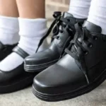 Close-up of a student wearing black leather lace-up shoes with white socks, with another pair in the background on a concrete floor.