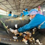 Workers in blue protective suits and masks sort trash on a moving conveyor inside a covered processing facility, picking litter from the belt.