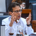 Man in glasses speaks at a meeting, hands raised as he explains, microphone in front of him