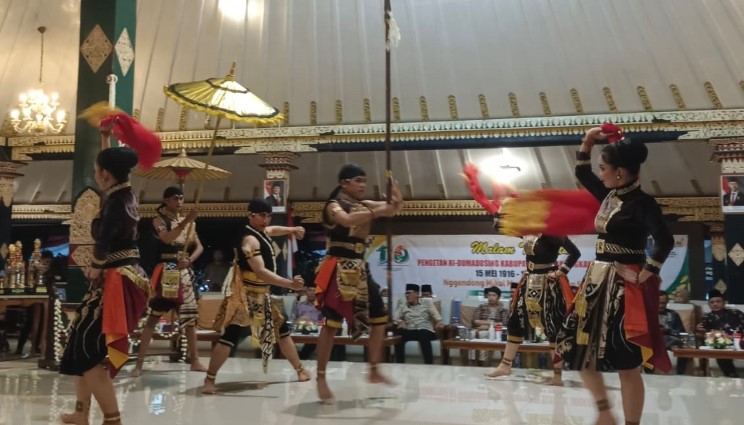 Group of Indonesian dancers in colorful costumes performing a traditional dance with fans and parasols indoors, on a polished stage.