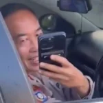 Officer in uniform taking a selfie with a cellphone inside a patrol car, reflected in the window panel.
