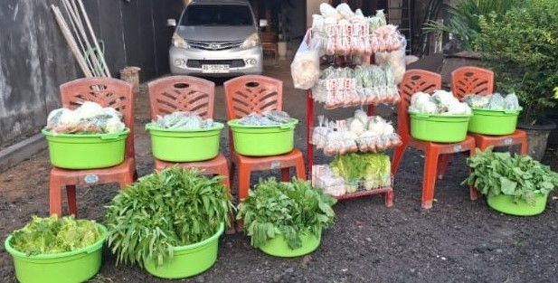 Street market stall with green buckets of leafy greens placed on orange plastic chairs and stacked snack bags in front of a parked car.