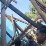 Group of people standing amid a collapsed timber frame with diagonal beams and a blue exterior wall behind them, inspecting the wreckage.
