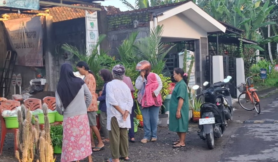 Group of people in hijabs and casual clothing stand near a street market stall with baskets and plants outside a small building; a motorcycle and bicycles are parked nearby.