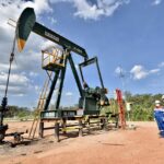 Oil pumpjack labeled Buraka in a dusty lot, with two workers in blue coveralls and hard hats nearby under a blue sky.