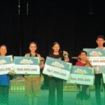 Group of children on a stage holding oversized ceremonial checks and trophies after a competition awards ceremony.
