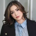 Portrait of a woman with shoulder-length wavy hair, wearing a light blue blouse and black blazer in an indoor setting.